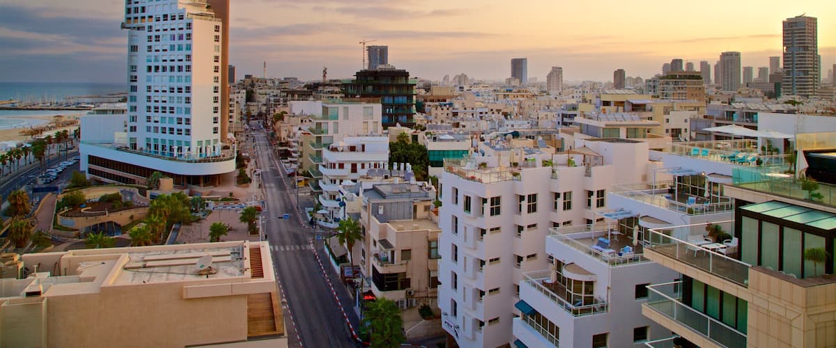 A cityscape with a prominent white building with balconies, a beach in the background, and a sunset sky.