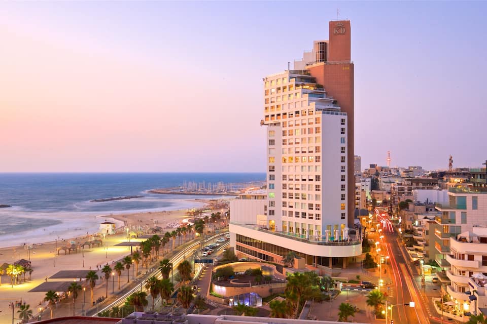 Un paisaje urbano costero con un hotel destacado, una playa y una calle concurrida al anochecer.