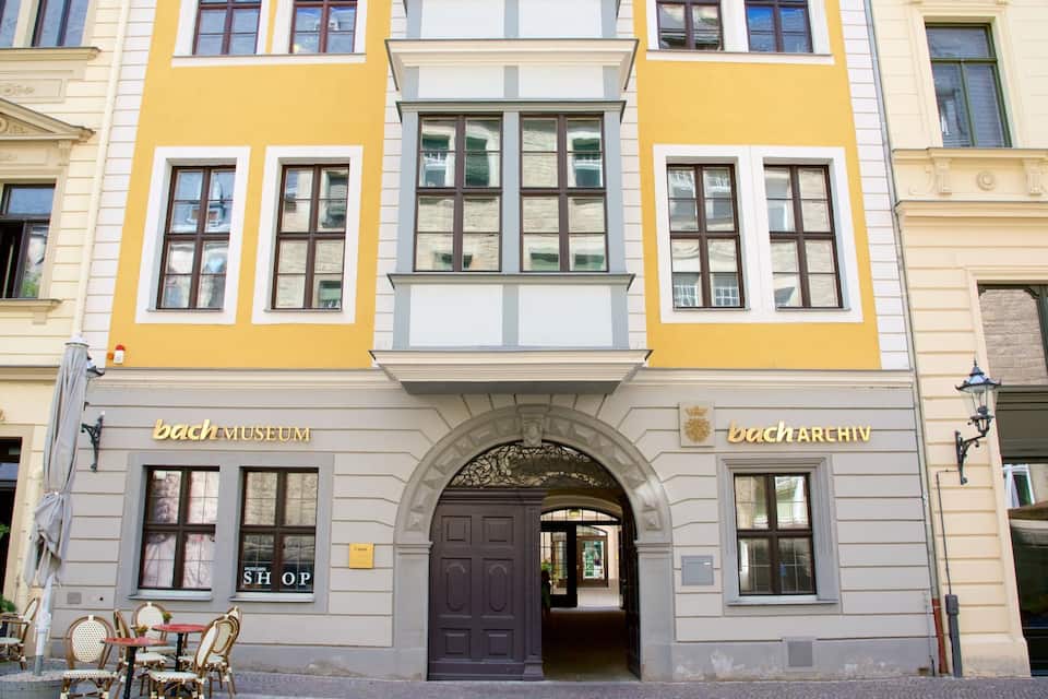 Um edifício com fachada amarela, duas entradas identificadas como 'bach Museum' e 'bach Archiv', e uma loja chamada 'Museums Shop' à esquerda.