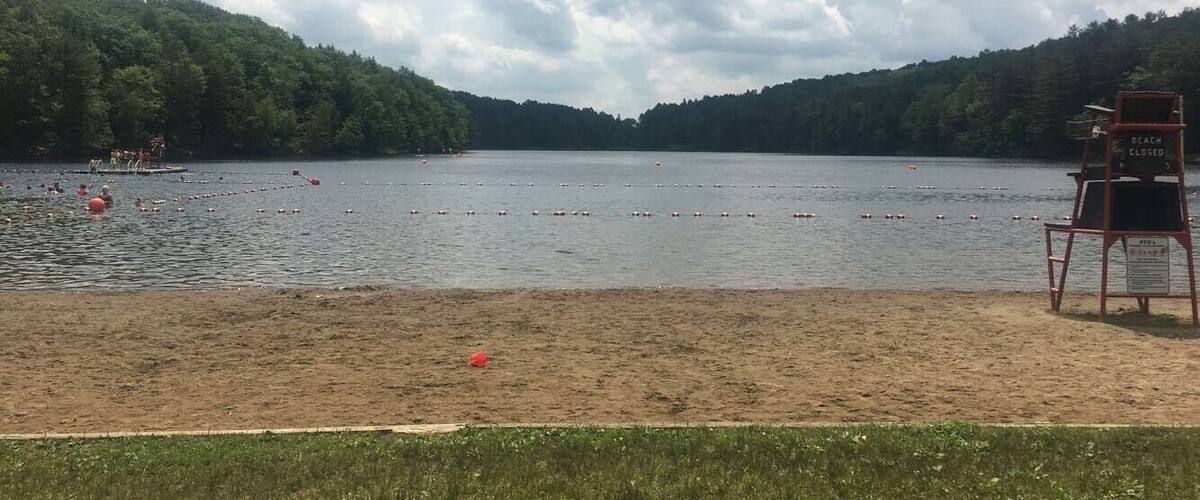 A lake with a beach chair and a 'BEACH CLOSED' sign, surrounded by forested hills.