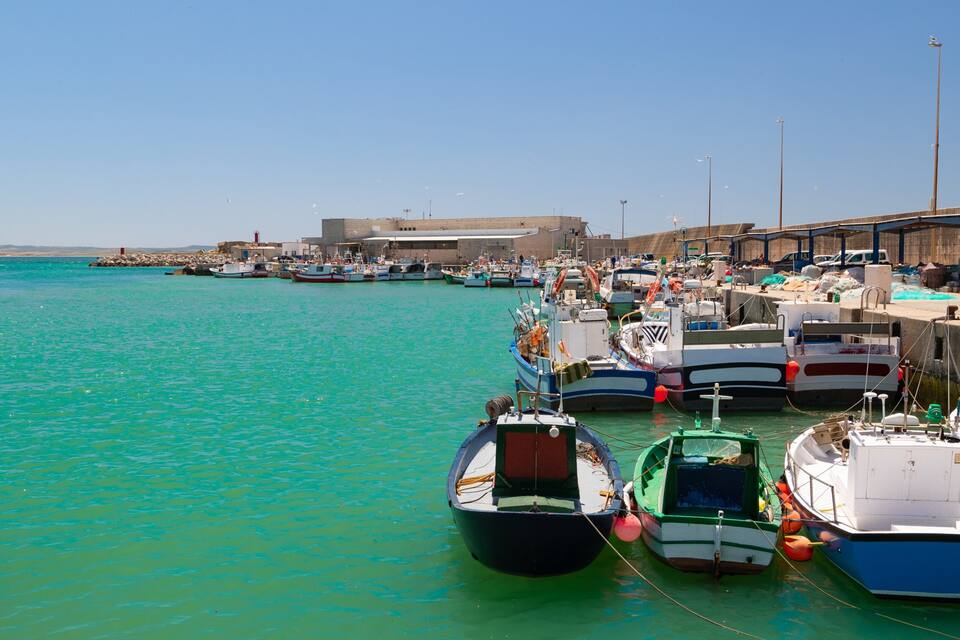 A harbor with numerous boats docked, a lighthouse, and a building in the background.