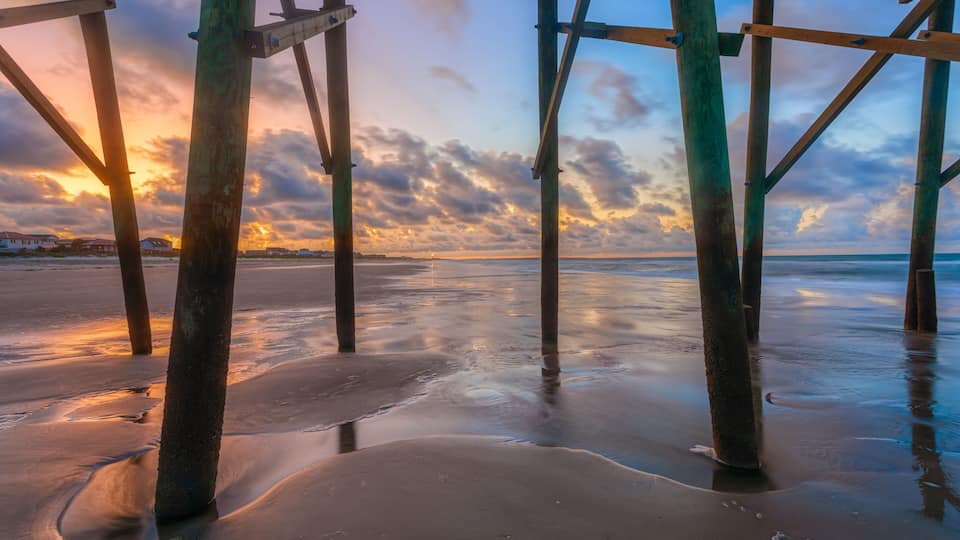 A wooden pier structure over a beach with a sunset in the background.