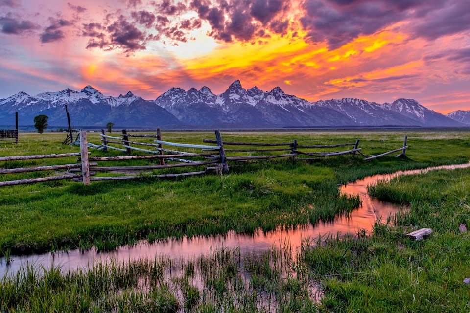 A sunset over a mountain range with a wooden fence and a small stream.