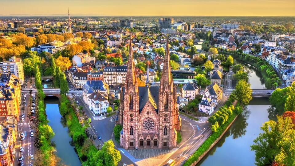 A cathedral with twin spires, surrounded by a river, trees, and urban buildings.