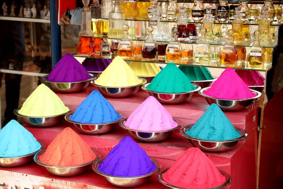 A display of colorful powders in metal bowls, likely for a festival or celebration, with a background of various bottles and containers.