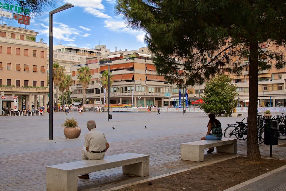 Uma praça com bancos, árvores e edifícios com placas como "Banco del Fucino".