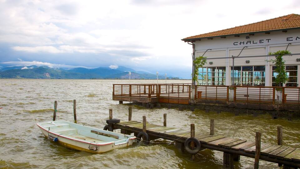 A wooden dock extending into a lake with a small boat tied to it. In the background, there is a building with the sign "CHALET EM" and mountains in the distance.