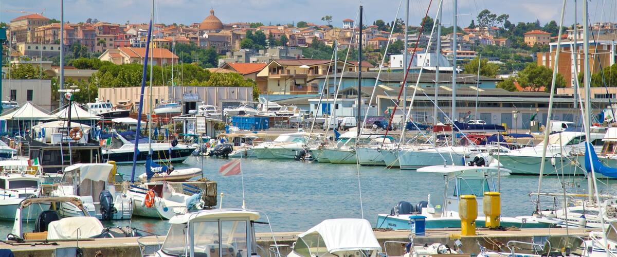 A marina with various boats docked, a cityscape in the background, and a prominent dome-shaped building.