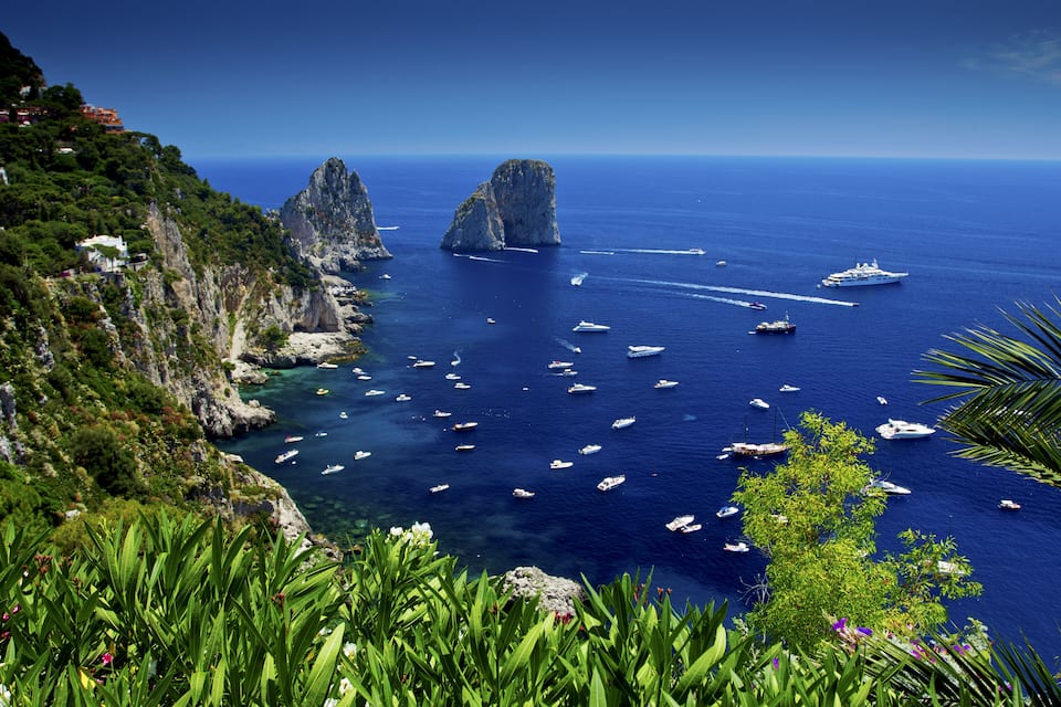 A coastal landscape with rocky cliffs, clear blue sea, and numerous boats.