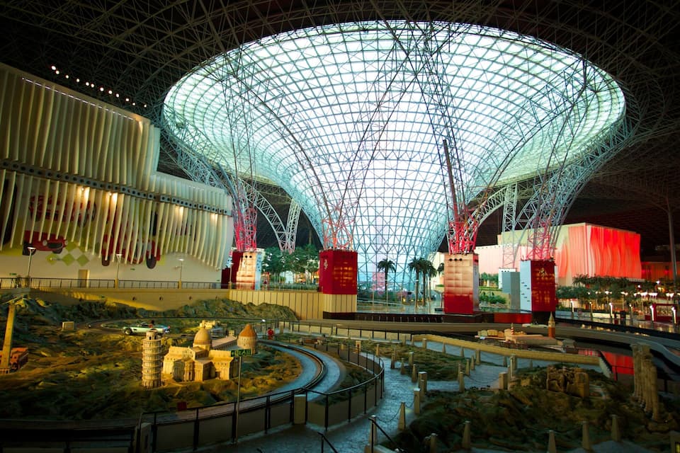 Uma grande estrutura em forma de cúpula iluminada, com um padrão semelhante a uma grade, cercada por uma área ajardinada com maquetes de edifícios e um rio.