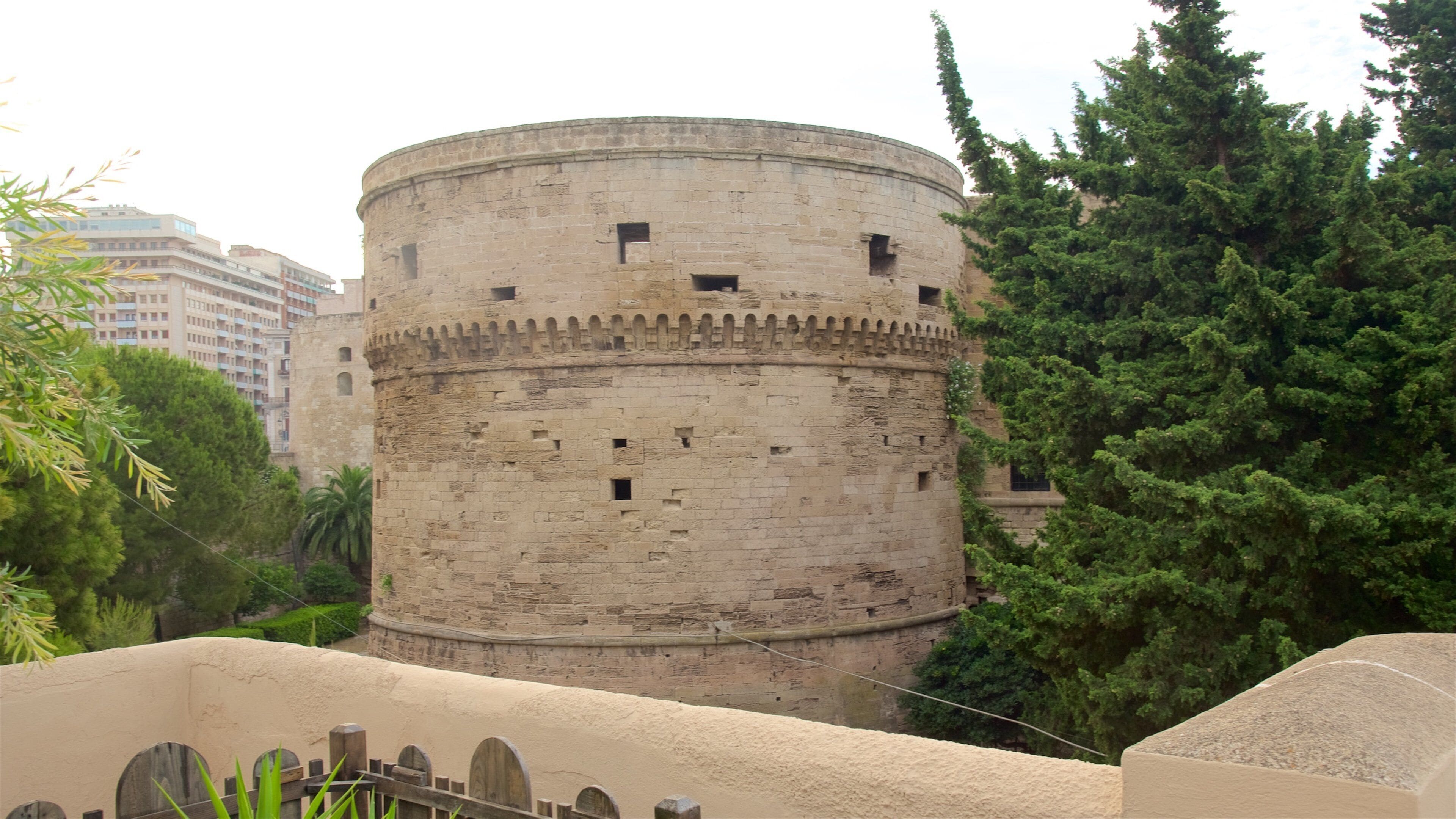 A historic stone tower with a crenellated top, surrounded by lush greenery and modern buildings in the background.