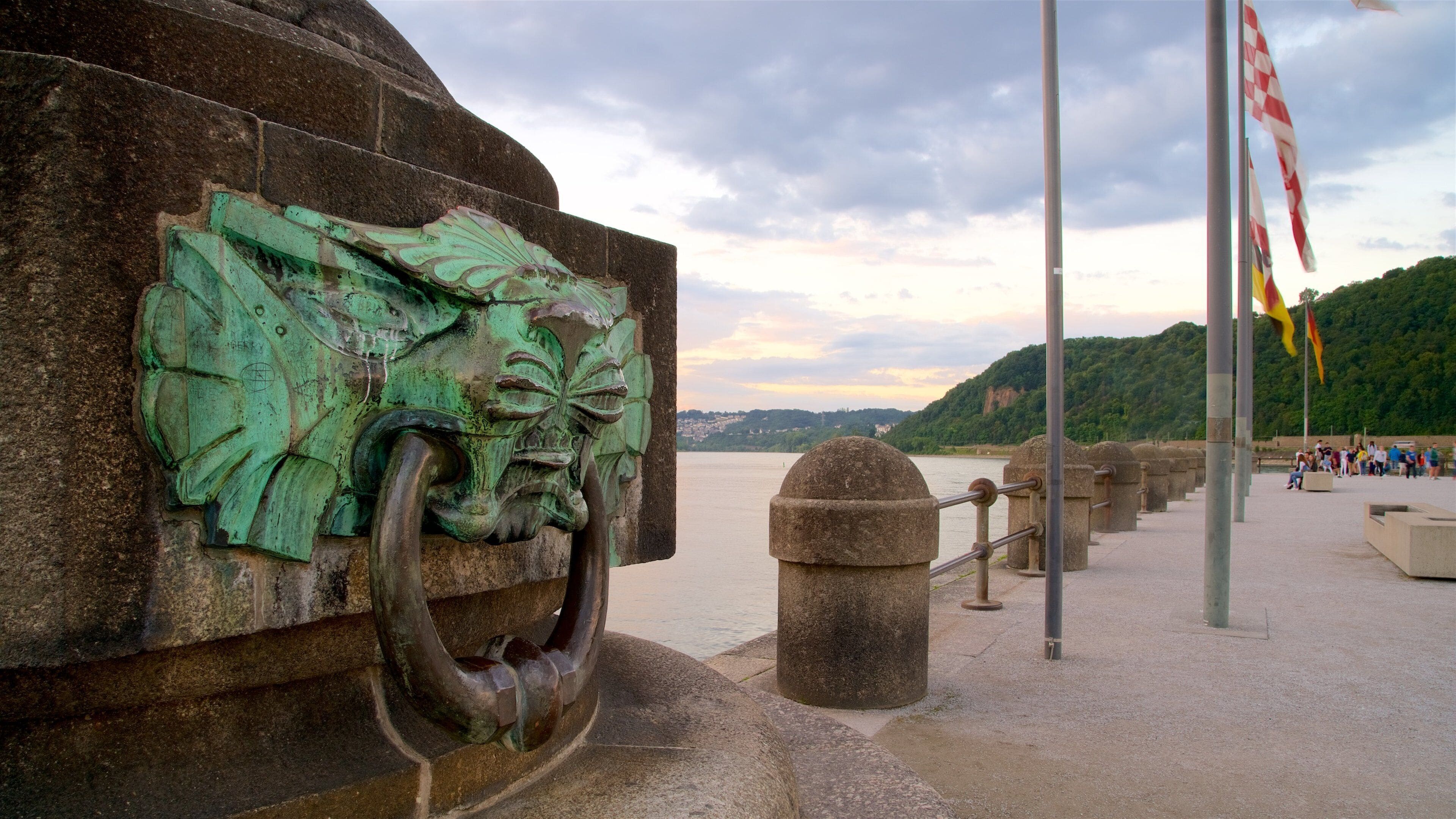 Un monument en pierre surmonté d’une sculpture métallique verdâtre, situé au bord de l’eau, et orné de drapeaux.