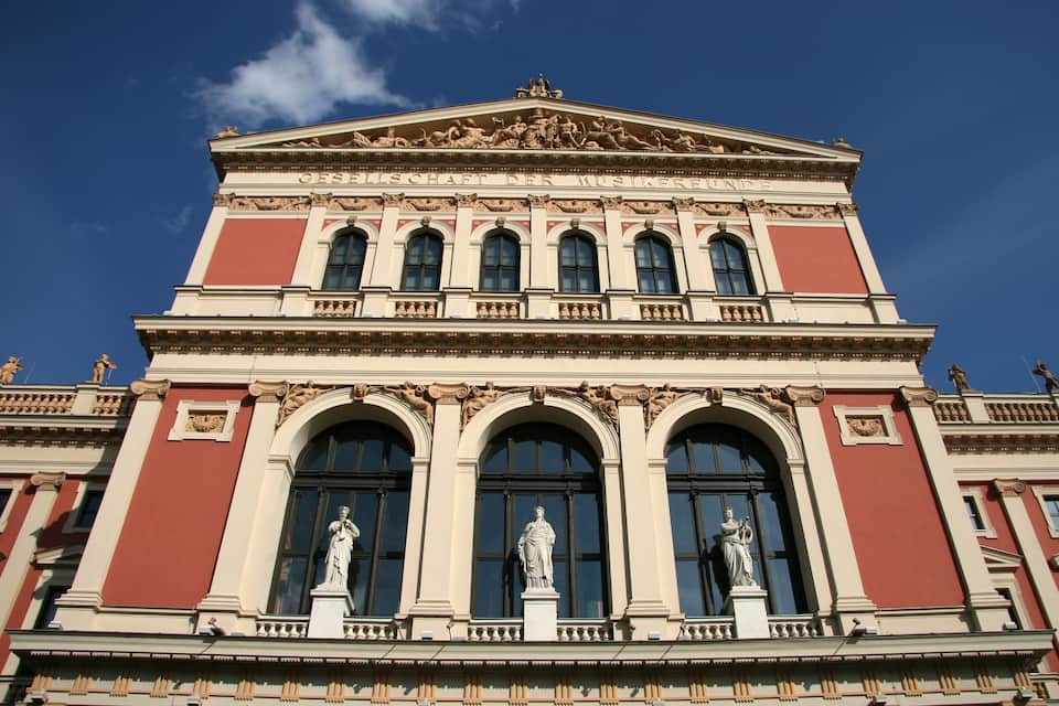 A classical building with a red facade, large arched windows, and statues on the ground floor.