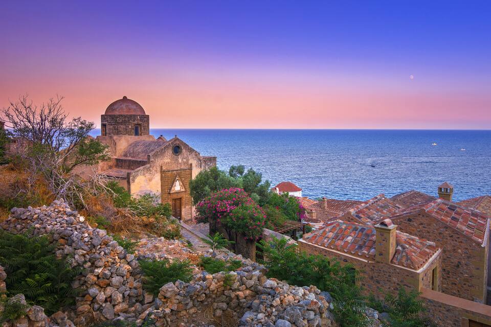 Un poble costaner amb edificis històrics, un mur de pedra i una vista del mar al capvespre.