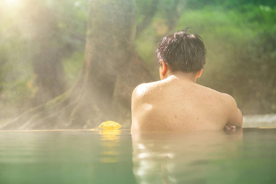 A person in a hot spring with water and steam rising, surrounded by greenery.