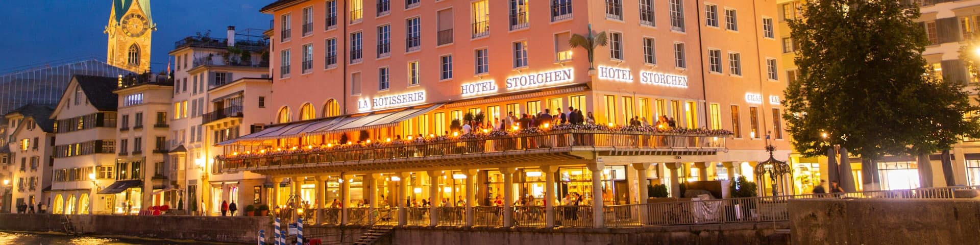 A riverside hotel with a clock tower and a church spire in the background.