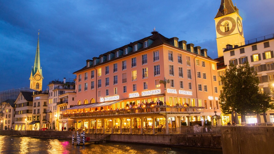 A riverside hotel with a clock tower and a church spire in the background.