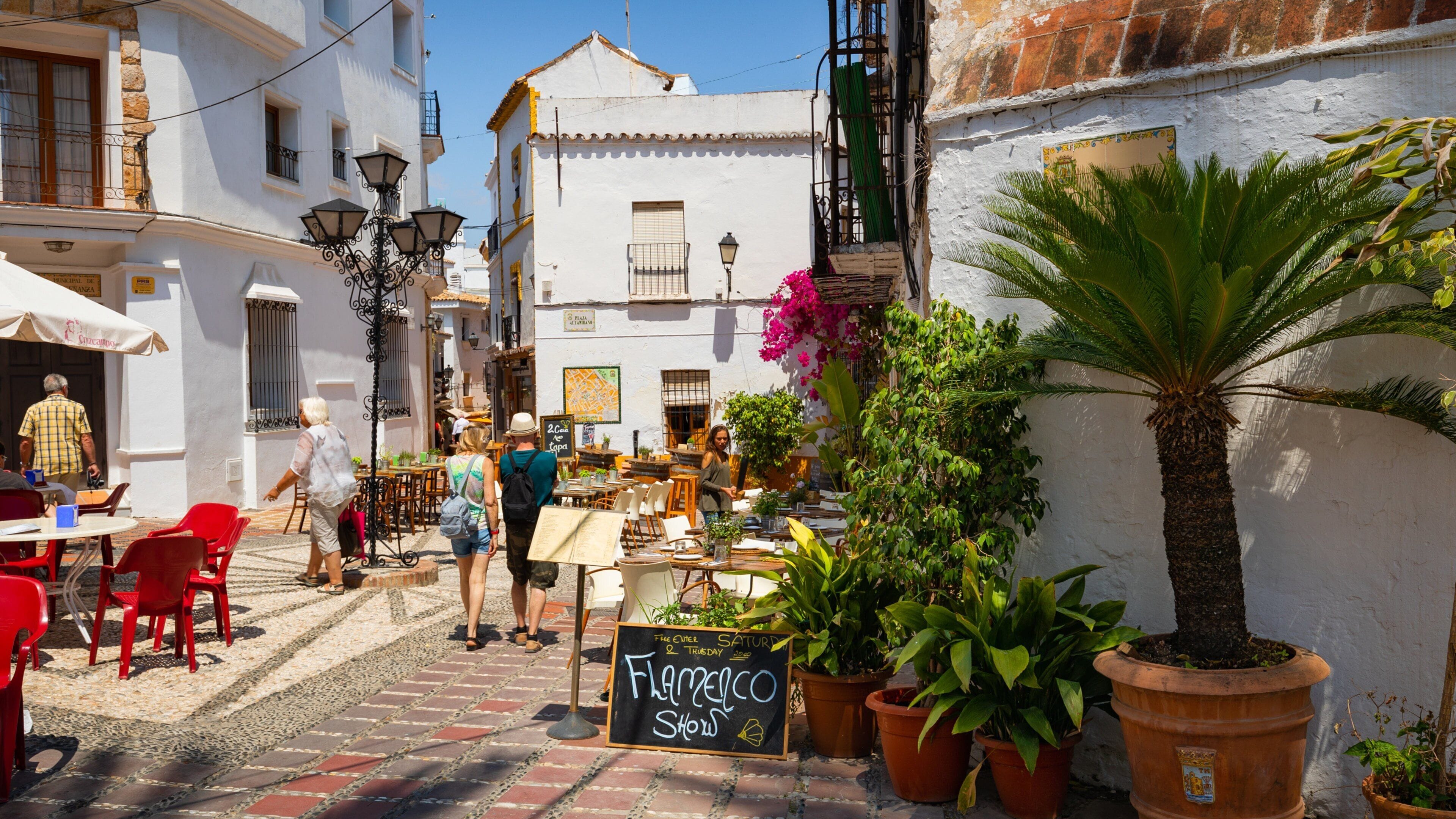 A cobblestone street with outdoor seating, potted plants, and a chalkboard sign advertising a show.