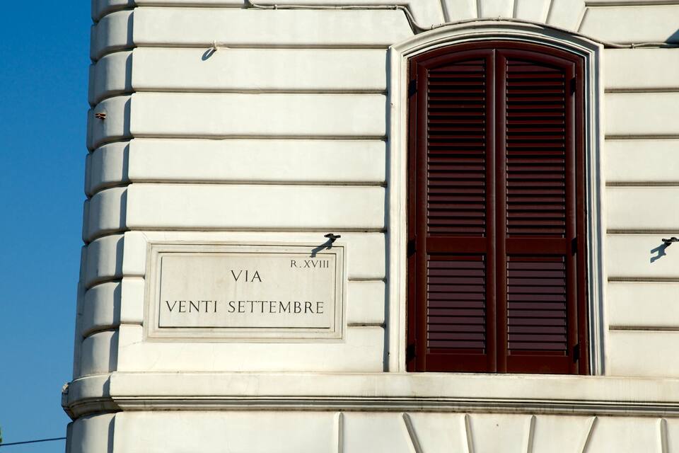 Facciata di un edificio con una targa che riporta “VIA VENTI SETTEMBRE” e una finestra con persiane in legno scuro.