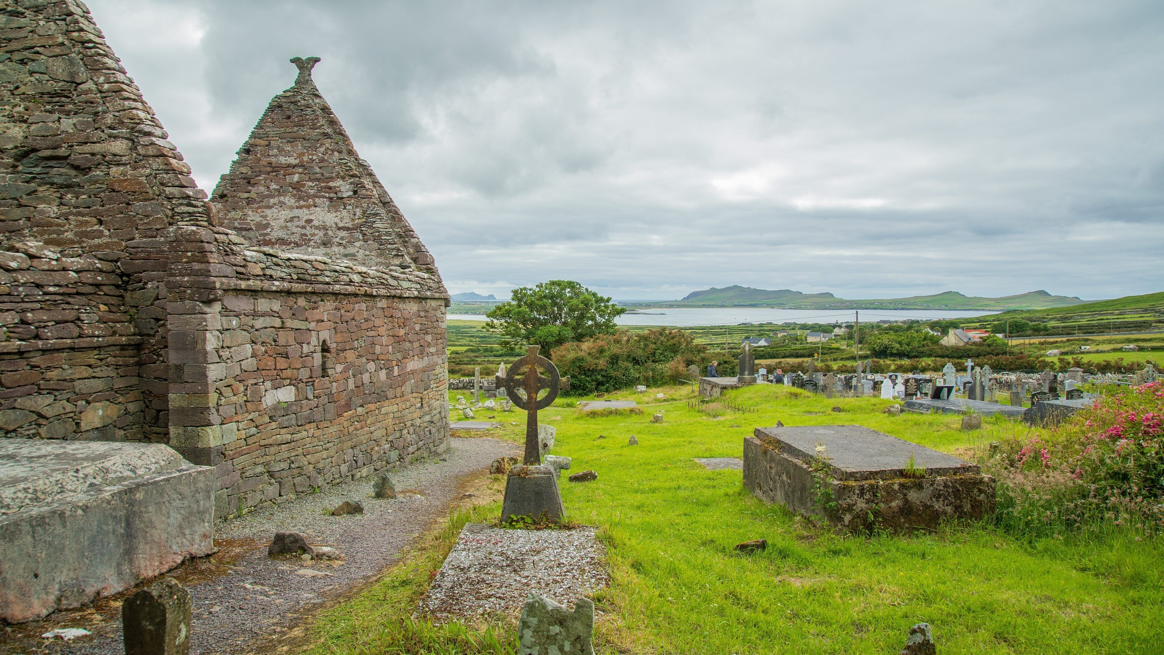 A stone church with a cross, a graveyard, and a view of the sea and hills in the background.