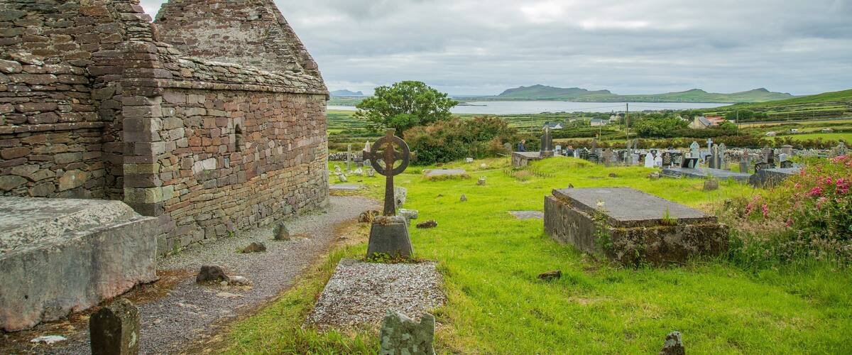 A stone church with a cross, a graveyard, and a view of the sea and hills in the background.