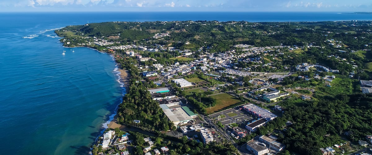 An aerial view of a coastal town with a beach, buildings, and a hilly landscape.