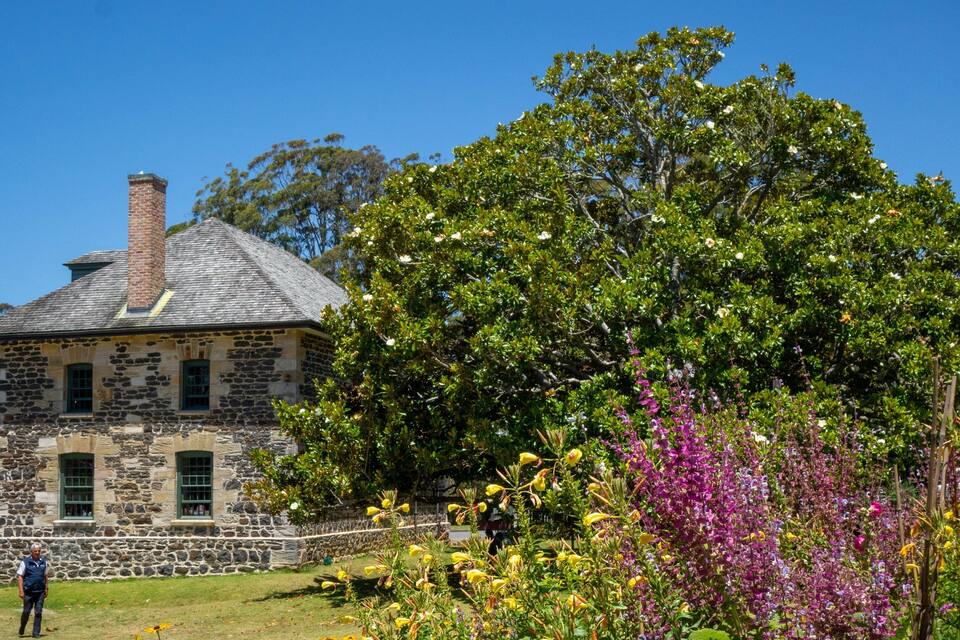 Une maison en pierre avec une cheminée, entourée d’une végétation luxuriante et de fleurs aux couleurs vives.