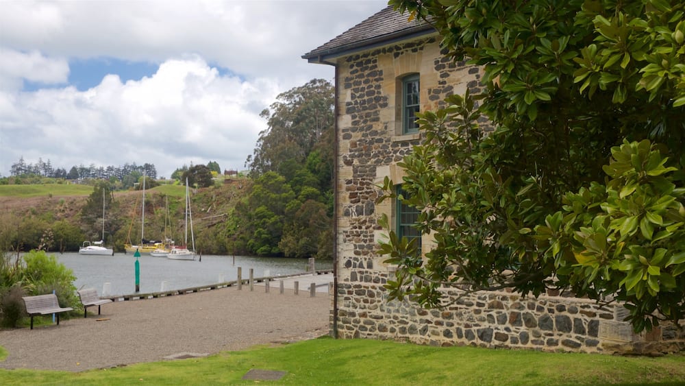 A stone building with a grassy foreground, a body of water with boats, and a hill with trees in the background.