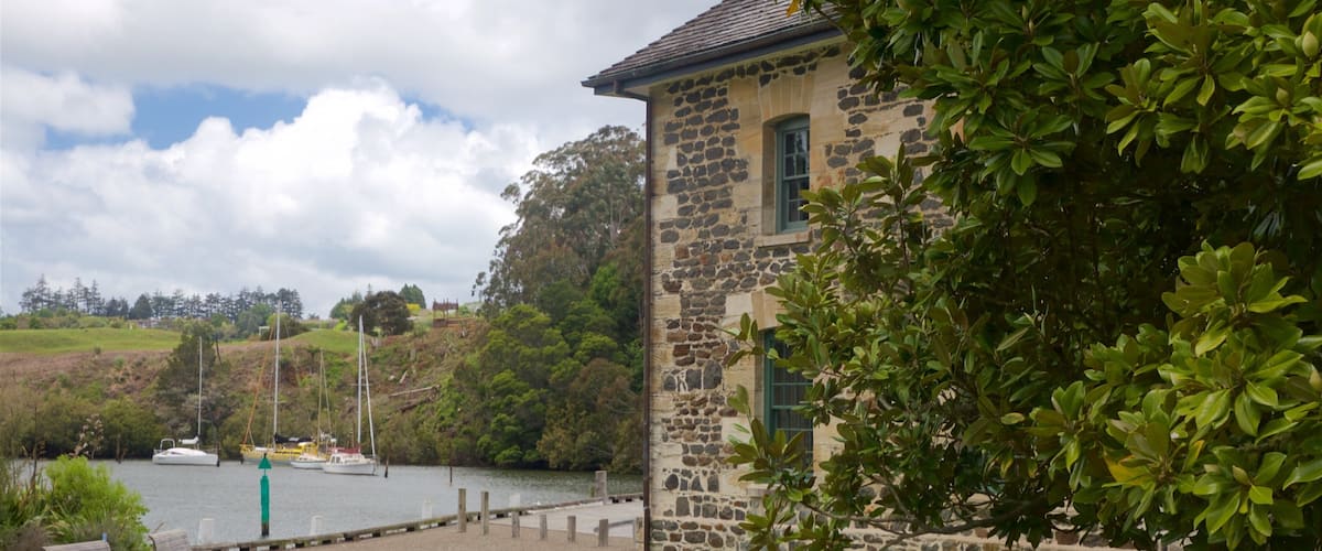 Ein Steinhaus mit einer grasbewachsenen Vorderseite, ein Gewässer mit Booten und ein Hügel mit Bäumen im Hintergrund.