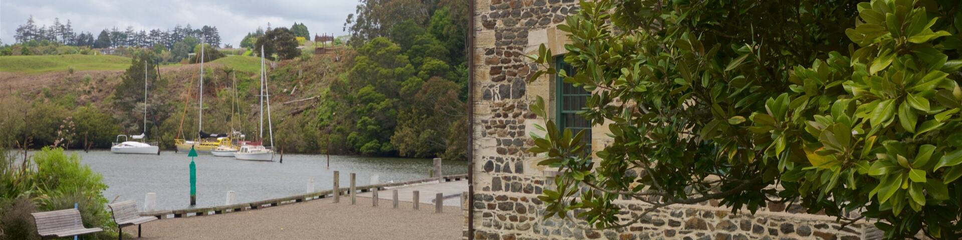 A stone building with a grassy foreground, a body of water with boats, and a hill with trees in the background.