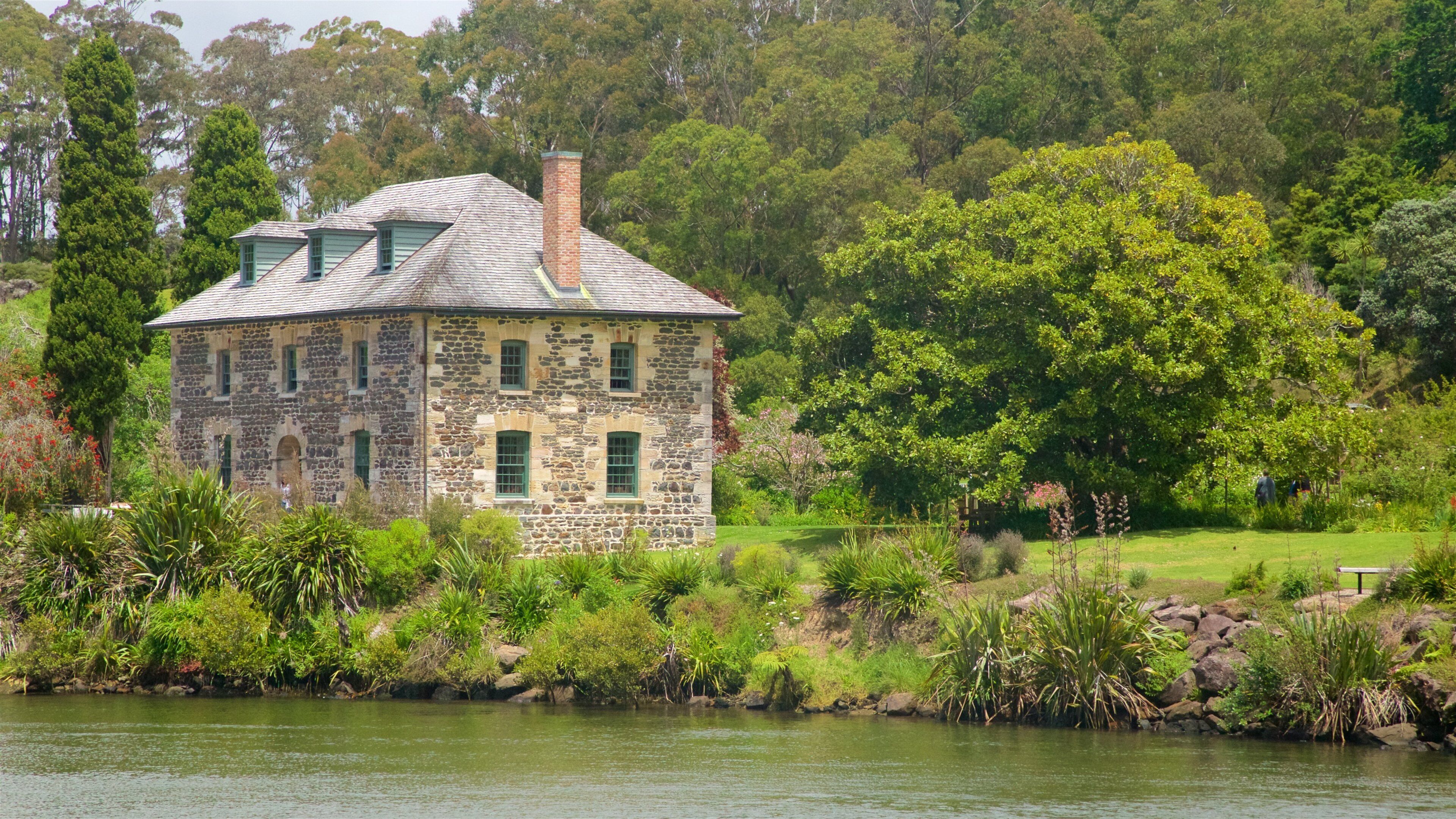 A stone house with a green roof by a lake, surrounded by lush greenery and trees.