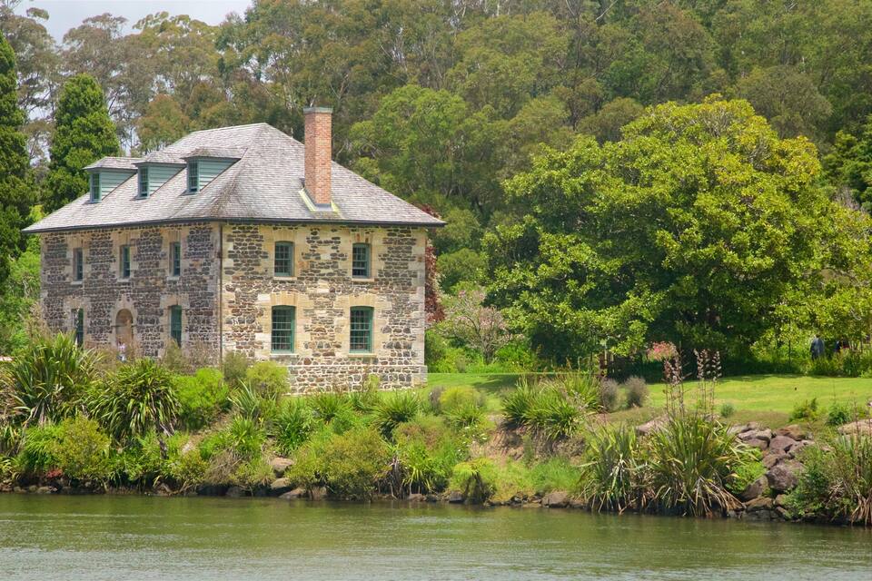 A stone house with a green roof by a lake, surrounded by lush greenery and trees.
