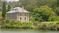 A stone house with a green roof by a lake, surrounded by lush greenery and trees.