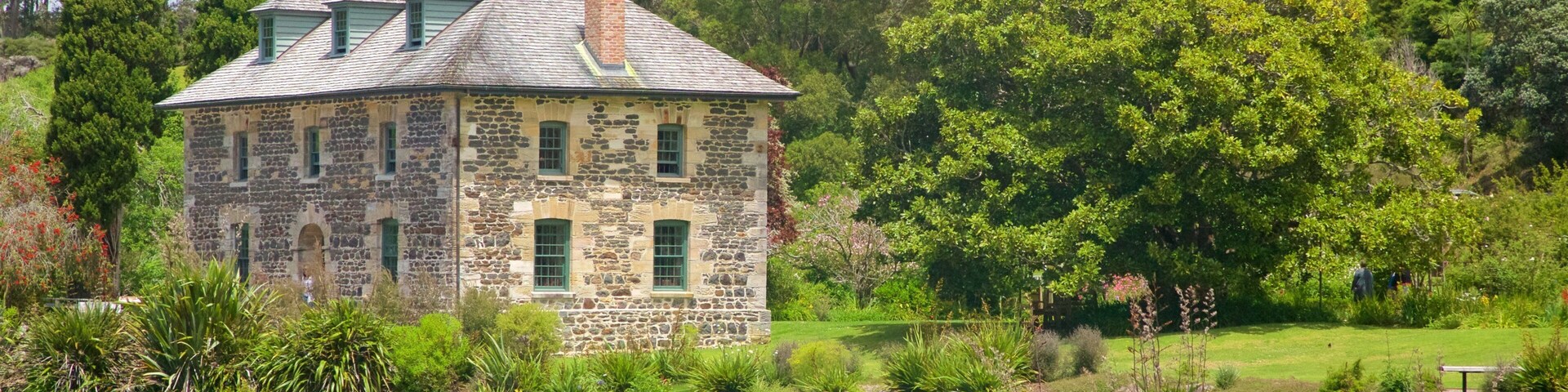 Una casa de piedra con techo verde junto a un lago, rodeada de exuberante vegetación y árboles.