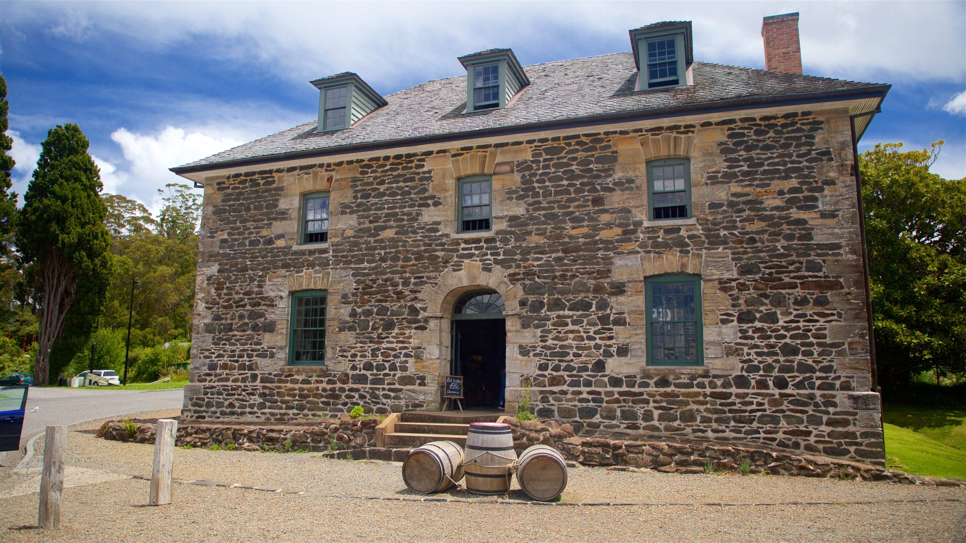 A stone building with a black door and green windows, surrounded by gravel and trees.