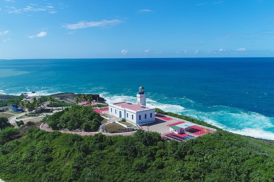A coastal lighthouse with a red and white building surrounded by lush greenery and the ocean.