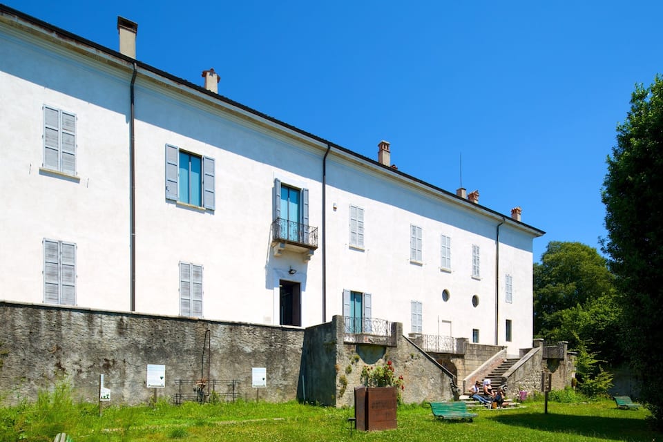 Um edifício branco com venezianas azuis, uma parede de pedra e um espaço gramado com bancos.