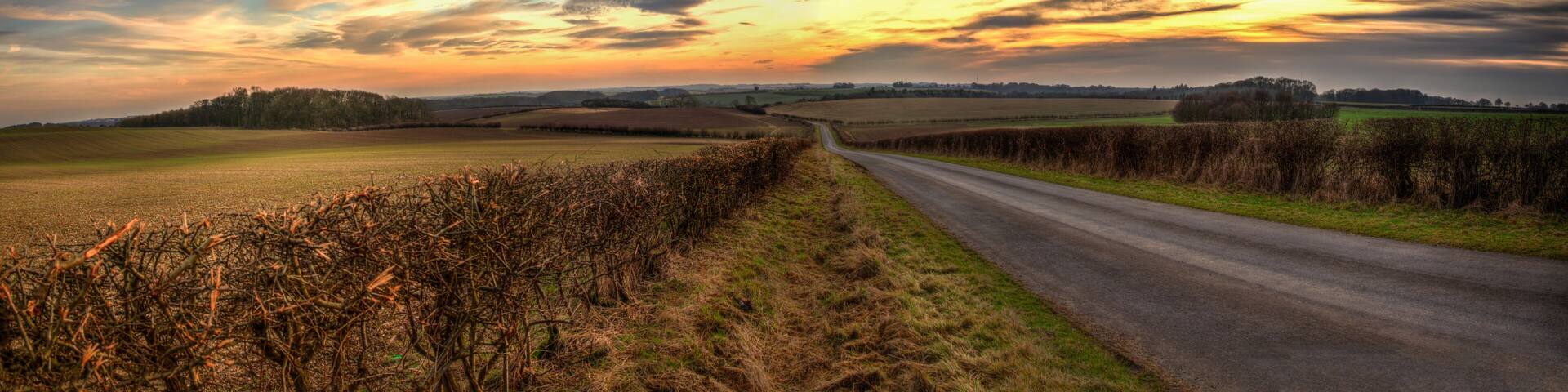 A rural landscape with a winding road, fields, and a dramatic sunset sky.
