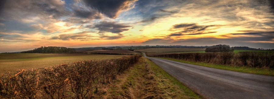 A rural landscape with a winding road, fields, and a dramatic sunset sky.