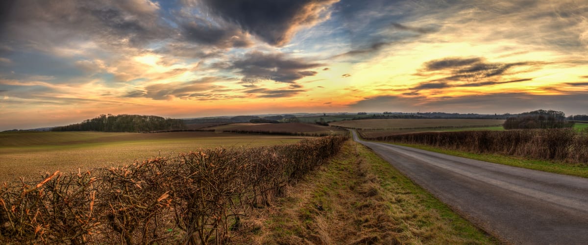 A rural landscape with a winding road, fields, and a dramatic sunset sky.