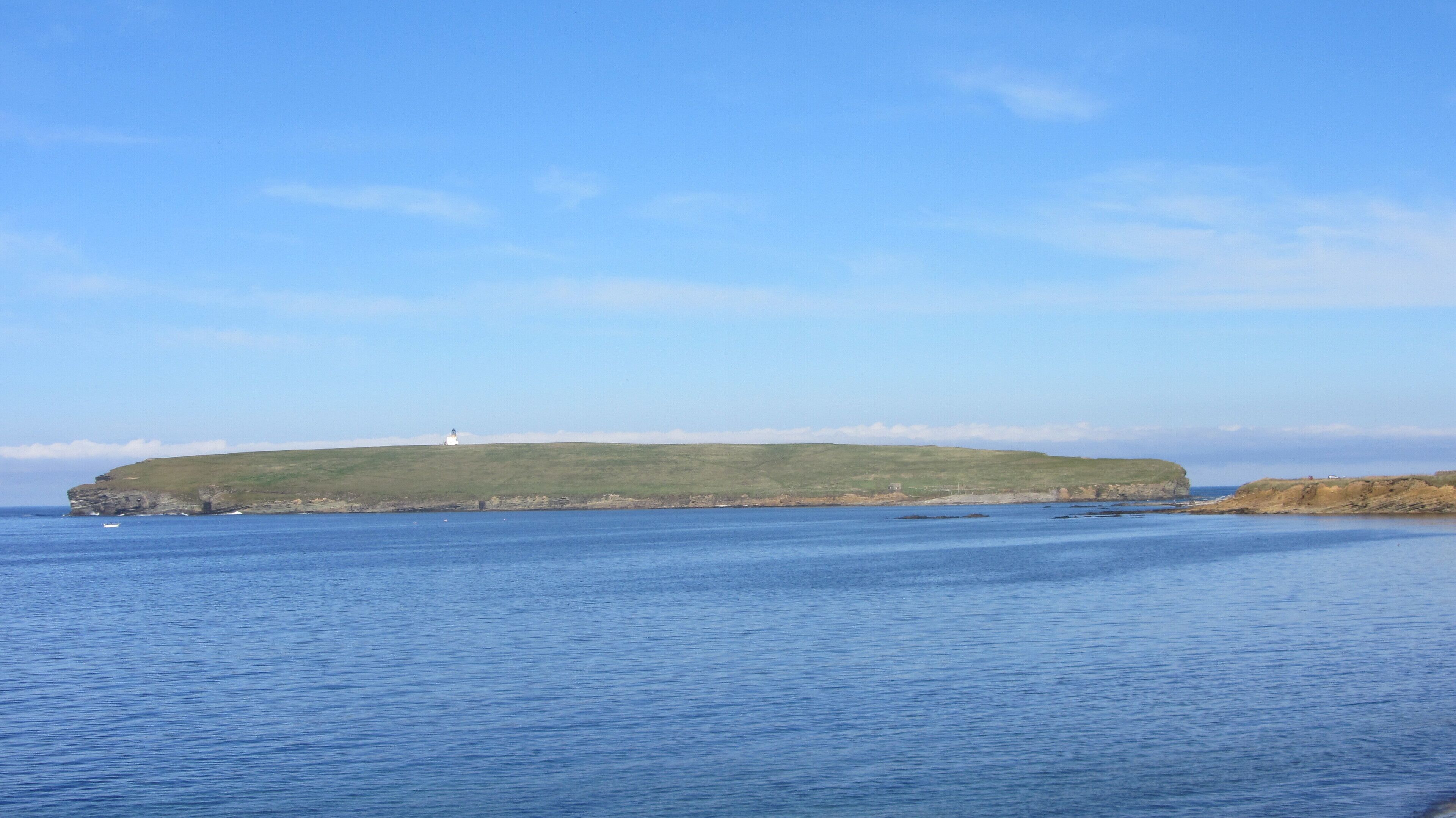 A coastal landscape with a lighthouse on a grassy hill, calm sea, and clear sky.