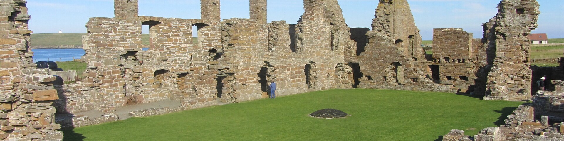 Las ruinas de un castillo medieval con un patio cubierto de césped, muros de piedra y un cielo despejado.