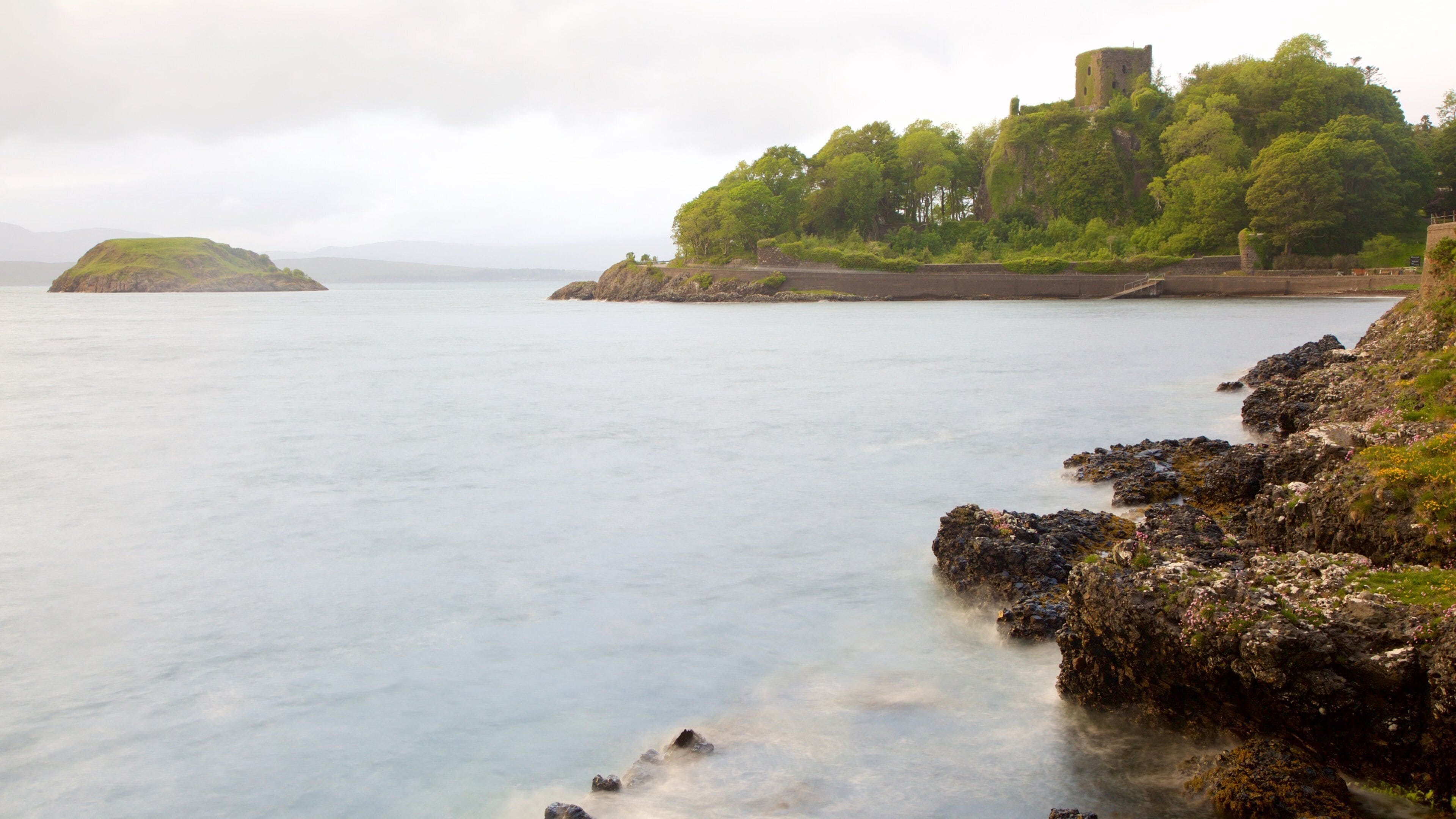Paisaje costero con un castillo en una colina, mar tranquilo, costa rocosa e isla distante.