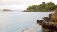 Un paysage côtier avec un château perché sur une colline, une mer calme, une côte rocheuse et une île lointaine.