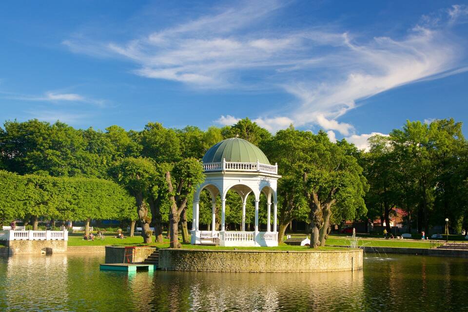 Un gazebo con cupola verde su un’isola di pietra in un parco, con alberi e cielo azzurro.