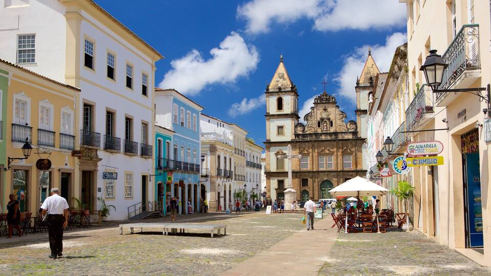 A cobblestone street lined with colorful buildings, a prominent church with a bell tower, and outdoor seating areas.