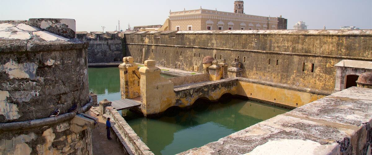 Un’antica fortezza con un ponte sopra un fossato, circondata da vecchie mura in pietra e un grande edificio sullo sfondo.
