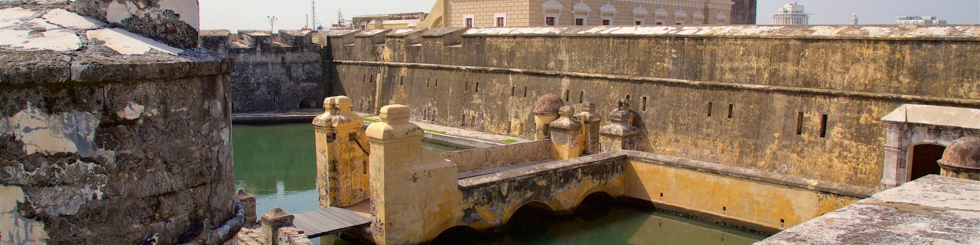 A historic fort with a bridge over a moat, surrounded by old stone walls and a large building in the background.