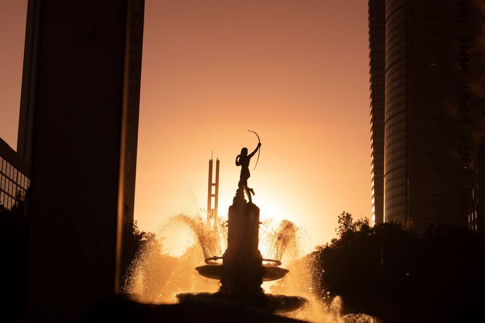 A silhouette of a statue of a figure with a bow and arrow in front of a sunset, with a bridge in the background.