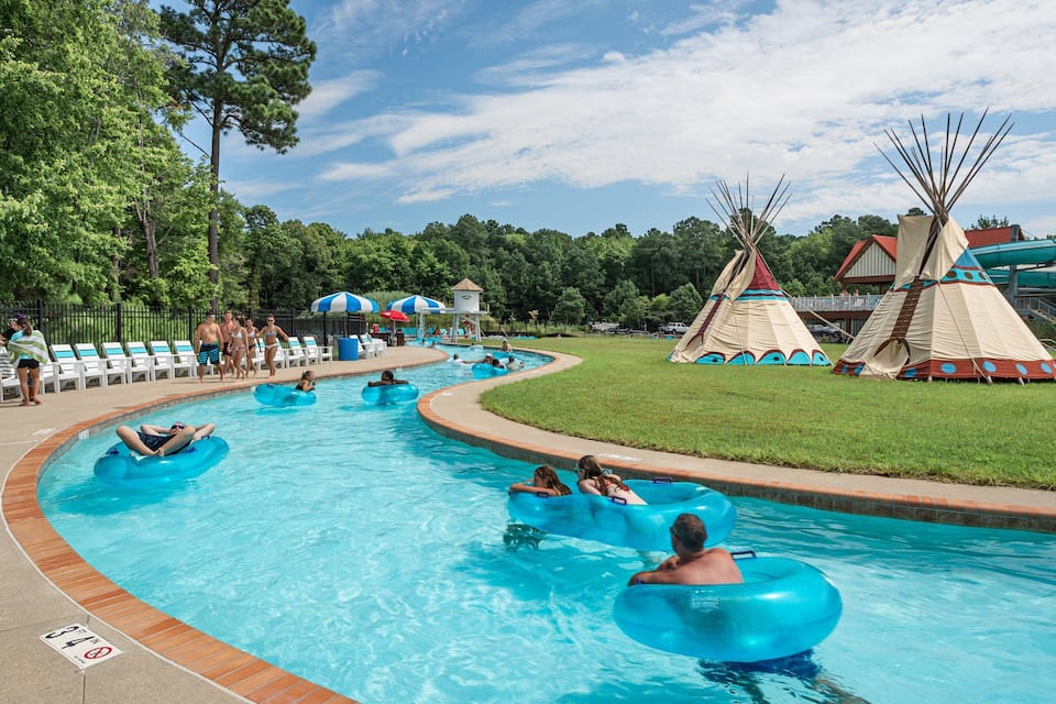 A swimming pool with people on inflatable rings, surrounded by a grassy area and teepees.